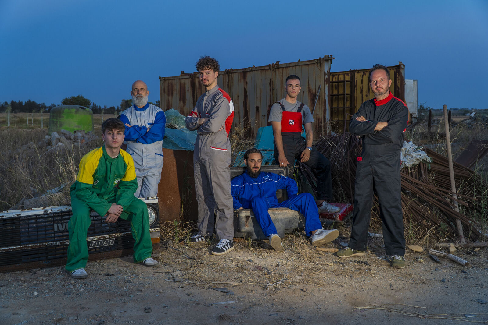 The six members of Kamafei standing among rusted metal containers and scrap at dusk, in colourful worksuits