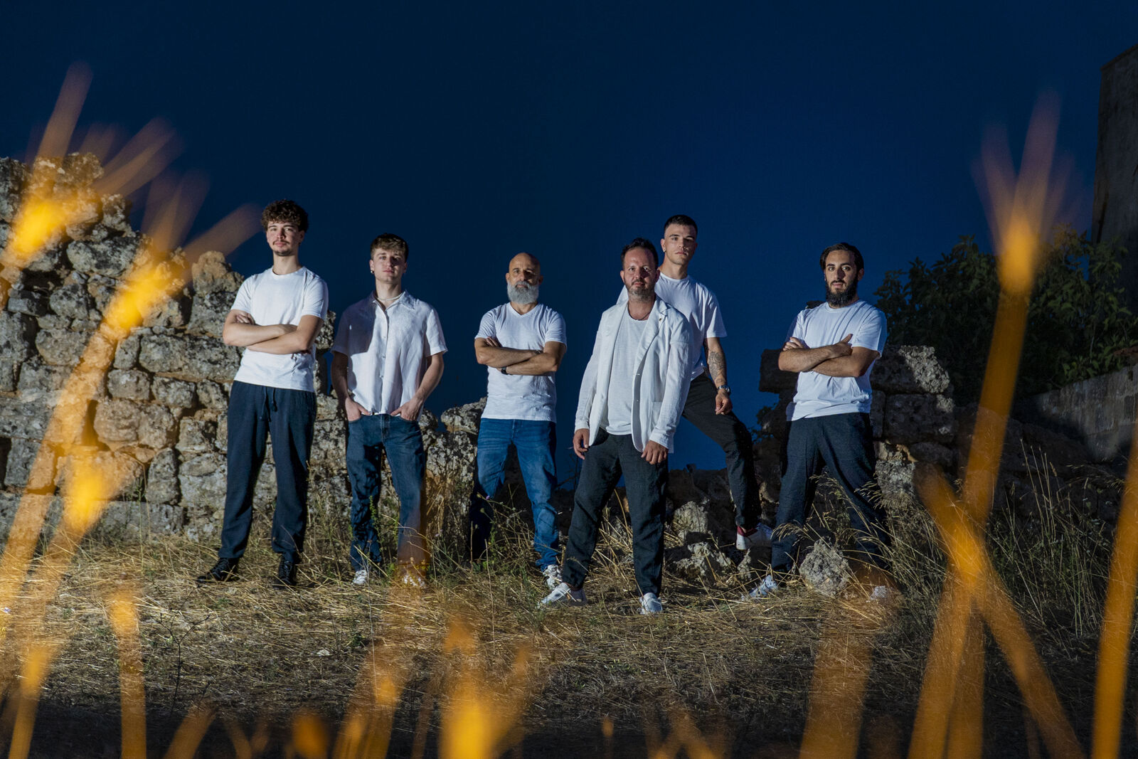 The band standing in white shirts among stone ruins at dusk, backlit by golden light through dry grass